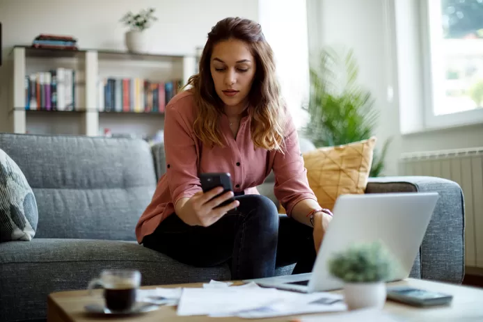 Vrouw zit in de zetel en zoekt op smartphone hoe ze haar maandelijkse factuur voor telecom kan verlagen