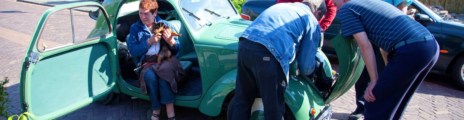 Mannen checken automotor van groene oltimer terwijl vrouw met hond in de auto zit.