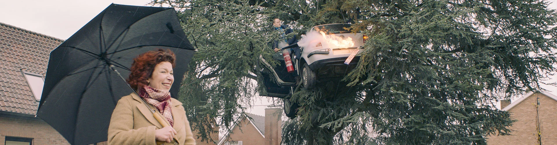 Vrouw staat met paraplu bij boom waar een brandende auto in vastzit