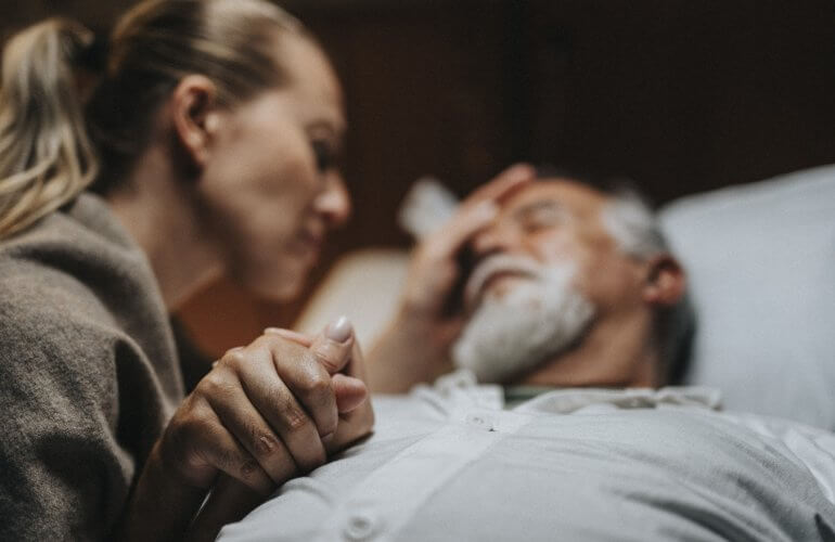 Vrouw houdt het hand van haar zwaarzieke man vast die op het bed ligt.